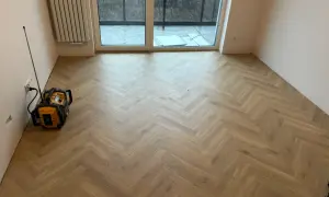 Nowe panele w jodełkę tworzą elegancką podłogę w pustym pokoju. Widać też grzejnik i drzwi tarasowe.
