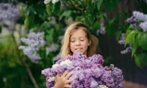 Uśmiechnięta dziewczynka z zamkniętymi oczami trzyma bukiet bzu. Wokół kwitnie bez fioletowy, tworząc magiczną atmosferę.