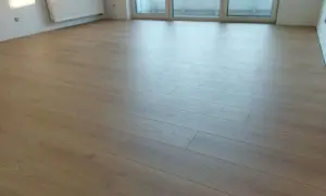 Jasne panele ułożone wzdłuż przedpokoju, tworząc wrażenie przestrzeni. Widoczny grzejnik i drzwi balkonowe.