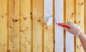 Ręka w rękawiczce maluje białą farbą drewniane panele ścienne wałkiem. Tak, można pomalować panele ścienne.