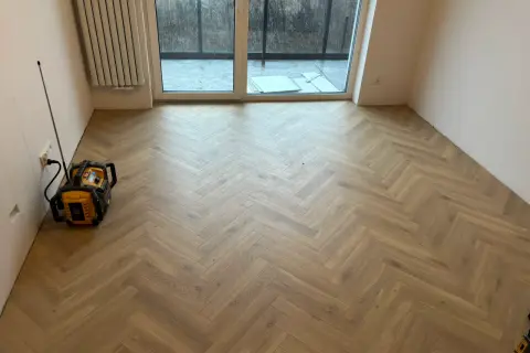 Nowe panele w jodełkę tworzą elegancką podłogę w pustym pokoju. Widać też grzejnik i drzwi tarasowe.