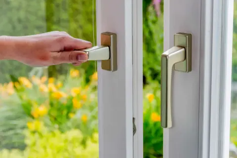 Dłoń chwyta klamkę okienną, pokazując jak wyjąć trzpień z klamki okiennej. W tle zielony ogród.