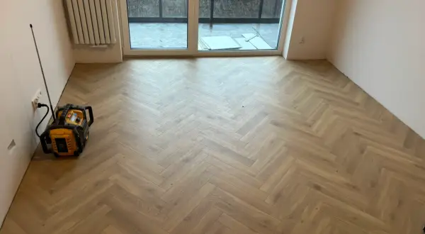 Nowe panele w jodełkę tworzą elegancką podłogę w pustym pokoju. Widać też grzejnik i drzwi tarasowe.