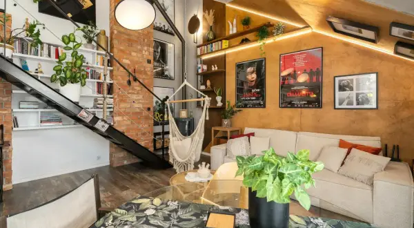 Salon w stylu loftowym z ceglaną ścianą, metalowymi schodami, wiszącym fotelem i wygodną kanapą.