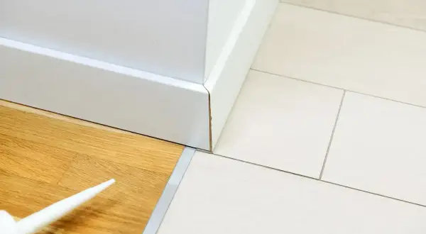 Biała listwa przypodłogowa łączy panele drewniane z płytkami. Widać tubkę z silikonem, gotową do pracy.