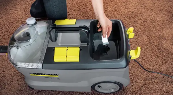 Ręka dodaje środek czyszczący do odkurzacza piorącego Karcher Puzzi 10/2. Dowiedz się, karcher rm 760 jak stosować, by skutecznie wyczyścić dywan.