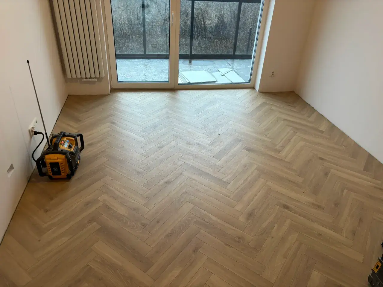 Nowe panele w jodełkę tworzą elegancką podłogę w pustym pokoju. Widać też grzejnik i drzwi tarasowe.