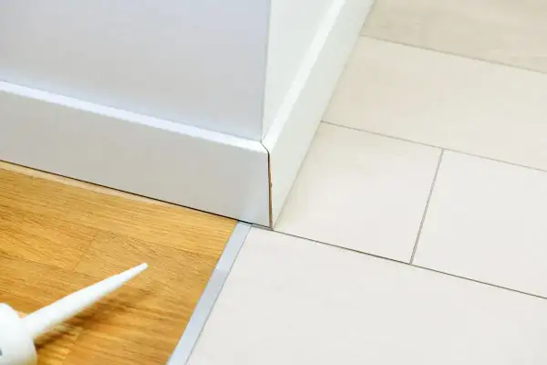 Biała listwa przypodłogowa łączy panele drewniane z płytkami. Widać tubkę z silikonem, gotową do pracy.