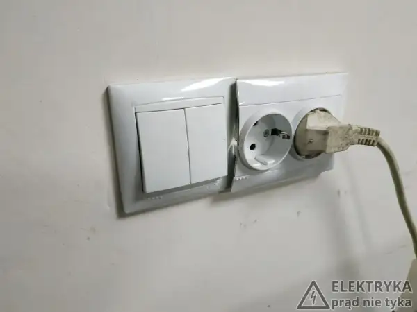 Biały montaż: włącznik światła i gniazdko elektryczne z wtyczką. Na ścianie widoczny znak ostrzegawczy "Elektryka prąd nie tyka".